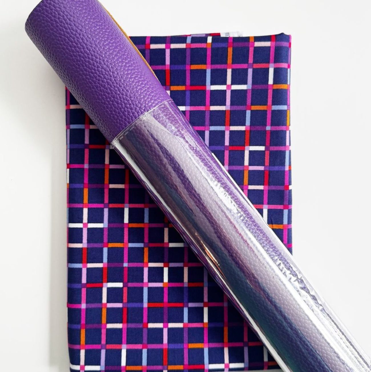 A fabric bundle featuring a folded purple cotton fabric with a colorful, modern geometric grid pattern, overlaid with a rolled piece of vibrant purple pebbled faux leather wrapped in clear plastic.