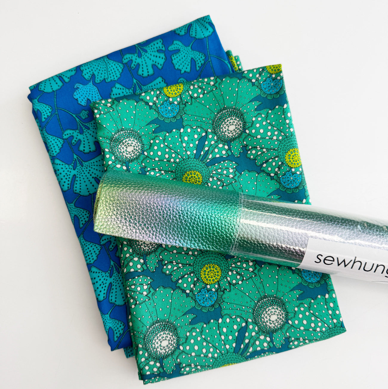 2-yard craft bundle featuring two folded cotton fabrics with teal and blue floral and ginkgo leaf prints, topped with a roll of green iridescent faux leather vinyl