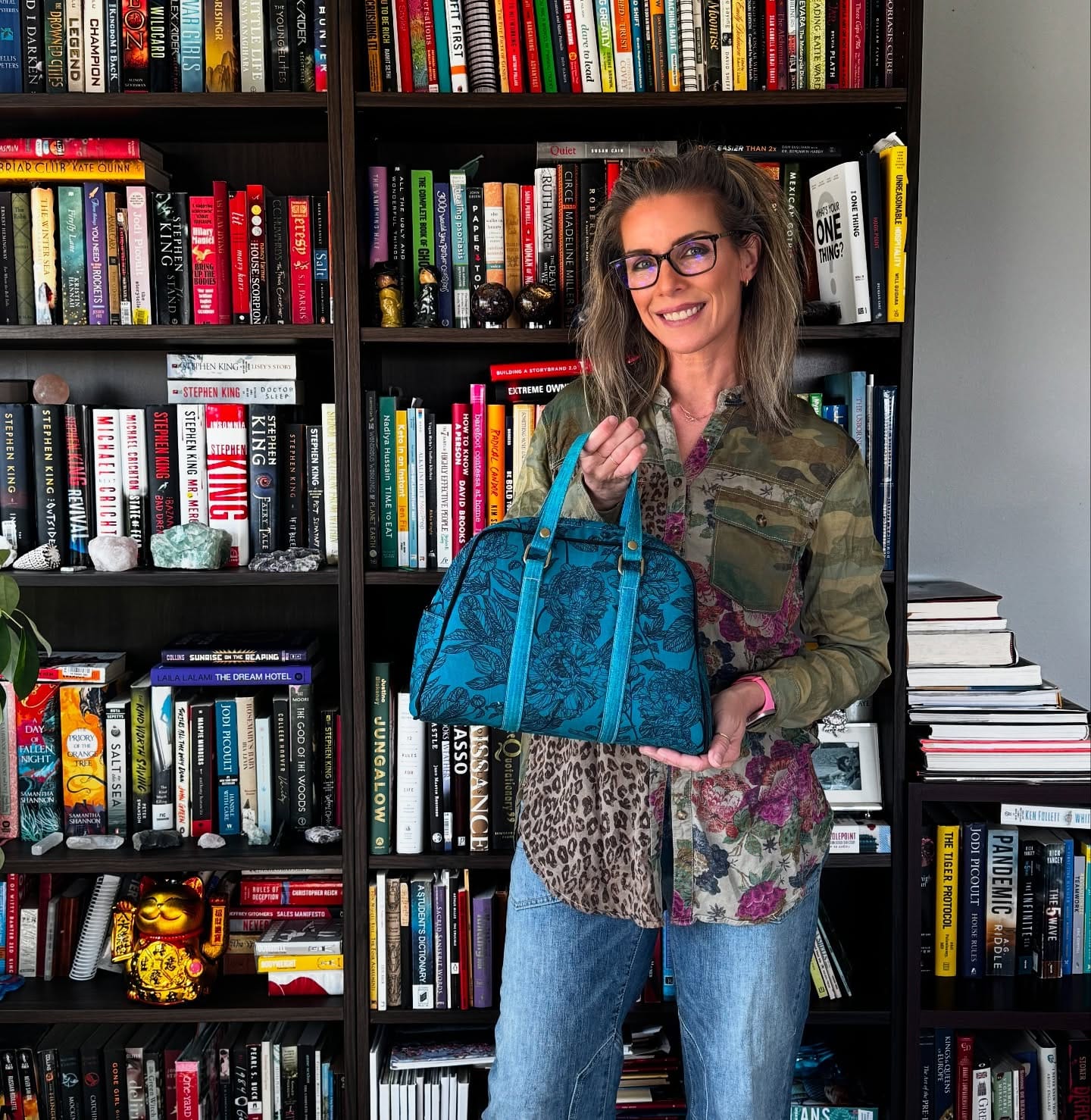 Woman with glasses holding a blue handbag in front of a bookcase.