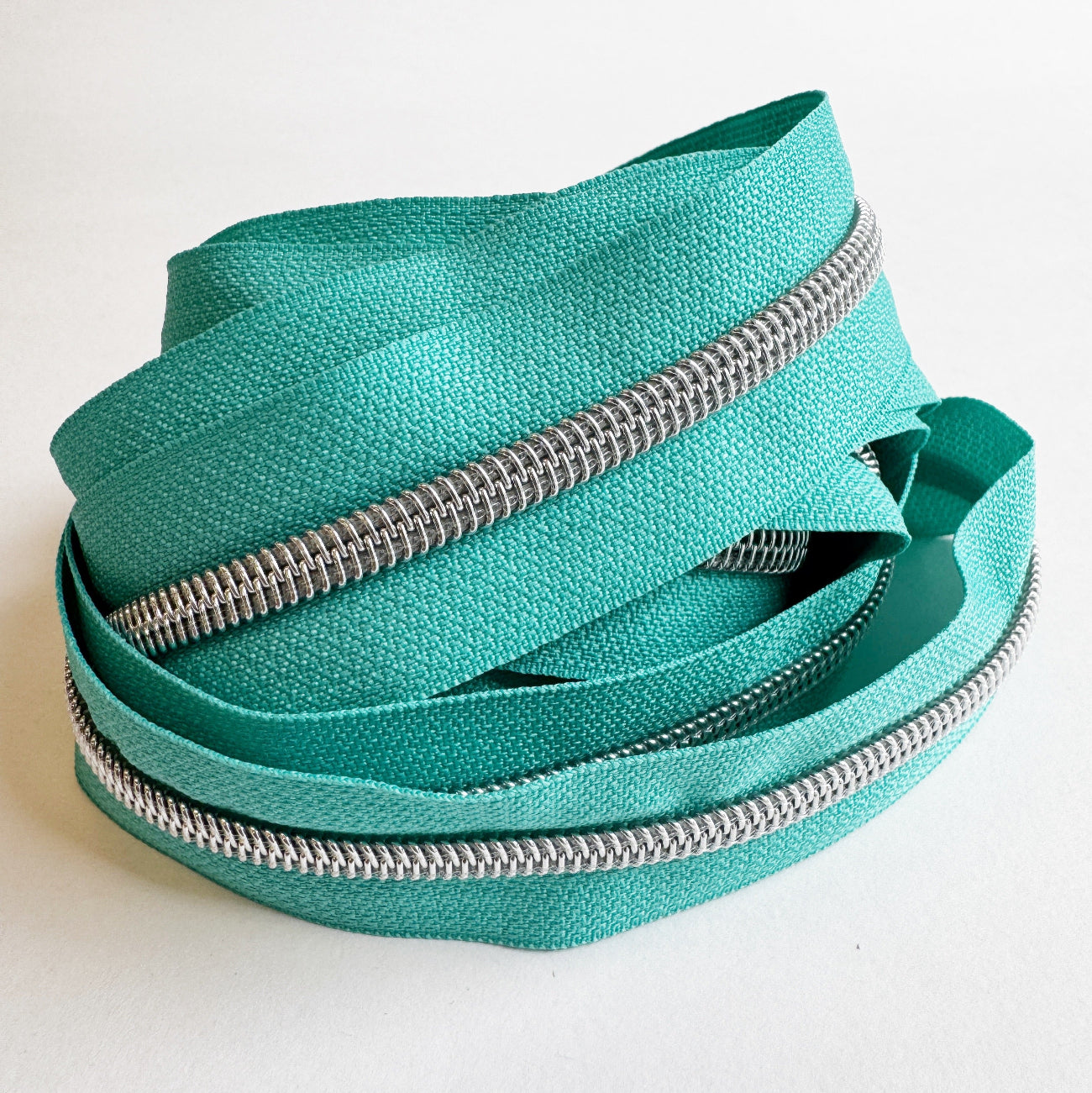 A length of teal nylon zipper tape loosely arranged into a circular, coiled nest shape on a clean white surface. The continuous rows of shiny silver zipper teeth track along the edges of the woven fabric, highlighting its flexibility.