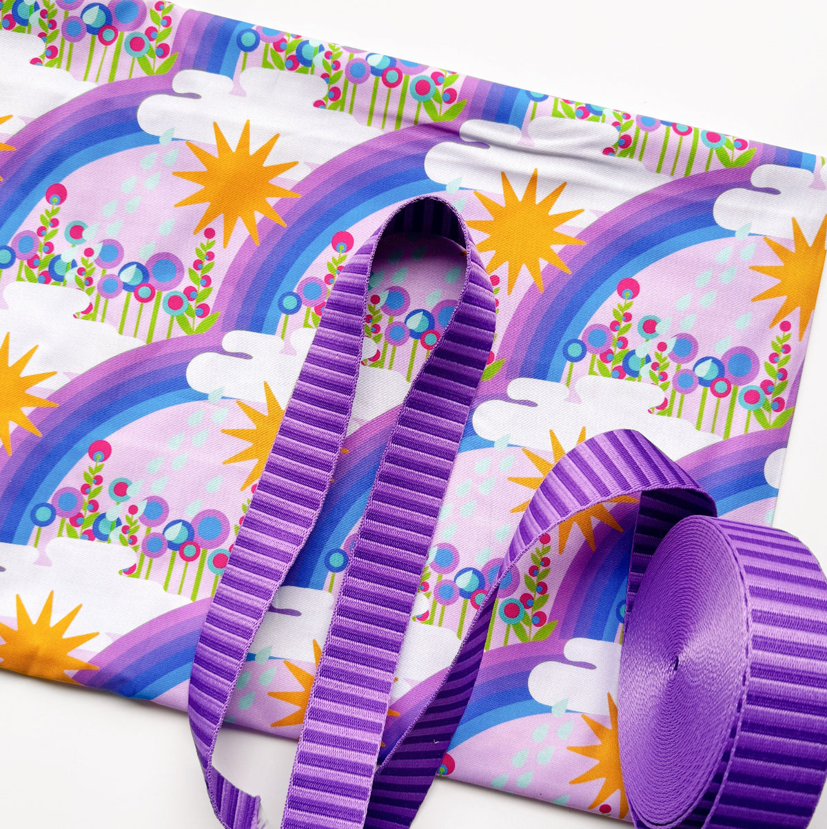 Light purple fabric with a colorful rainbow, sun, and cloud pattern paired with purple jacquard webbing. Postmark Lavender First Day Yardage by Alison Glass for Andover Fabrics.