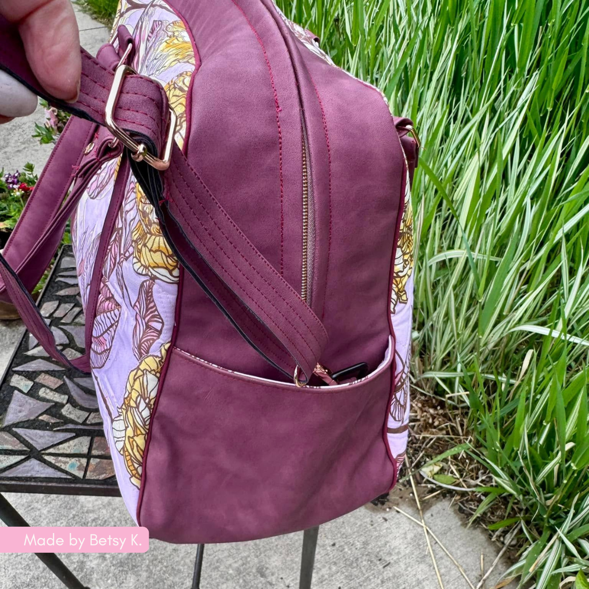 A side view of the magenta handbag with floral pattern panels, showing the zipper and side pocket details while positioned outdoors.