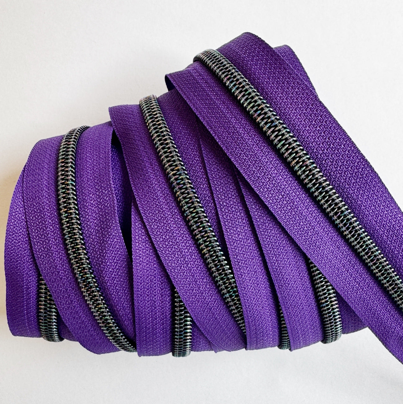 A close-up view of the purple zipper tape neatly layered and wrapped flatly to showcase several parallel rows of the metallic teeth. The shot emphasizes the rich texture of the purple woven fabric tape and the colorful iridescent shimmer on the gunmetal zipper teeth