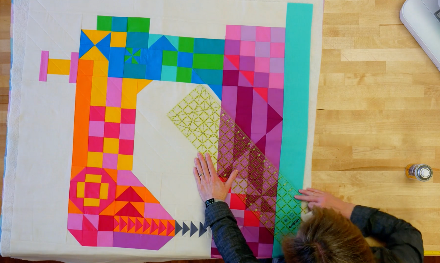 Natalie marking a colorful quilt with geometric blocks in the shape of a sewing machine on a wooden sewing table