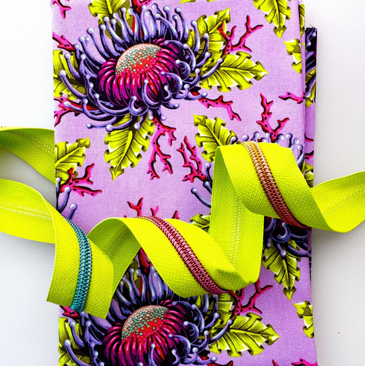 A folded fabric item, likely a napkin or small table linen, featuring a vibrant purple and lime green floral pattern with a bright neon yellow-green ribbon threaded through it, set against a plain white background.