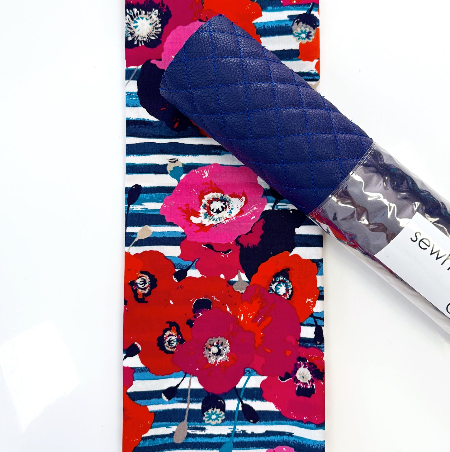 A folded fabric accessory with a navy quilted top and a colorful floral print featuring red, pink, and blue flowers on a striped background, shown next to a clear plastic tube labeled 'sewing'.