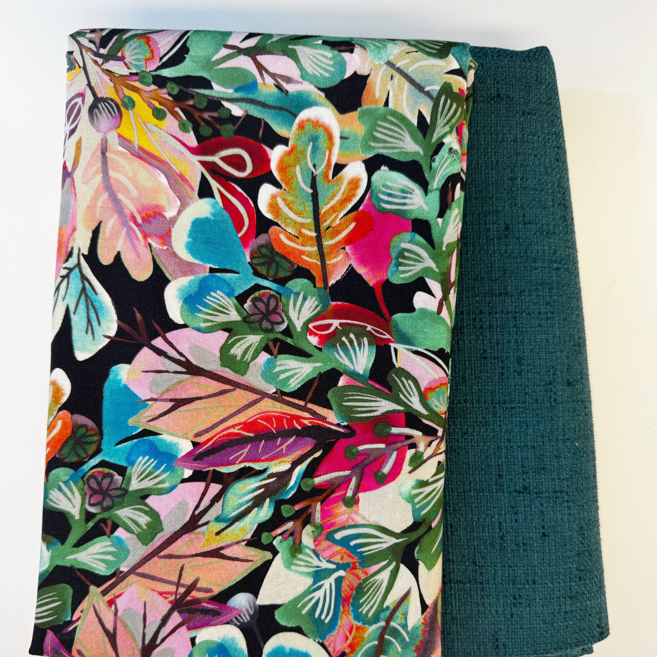 A coordinated fabric destash bundle featuring two neatly folded textiles against a white background. On the left is a vibrant denim fabric measuring 36 by 58 inches, printed with a dense, colorful tropical botanical pattern of leaves and abstract florals in shades of pink, teal, green, orange, and yellow on a black background. Peeking out from underneath on the right is a one-yard piece of textured, solid dark teal coordinating fabric with a woven, linen-like appearance.