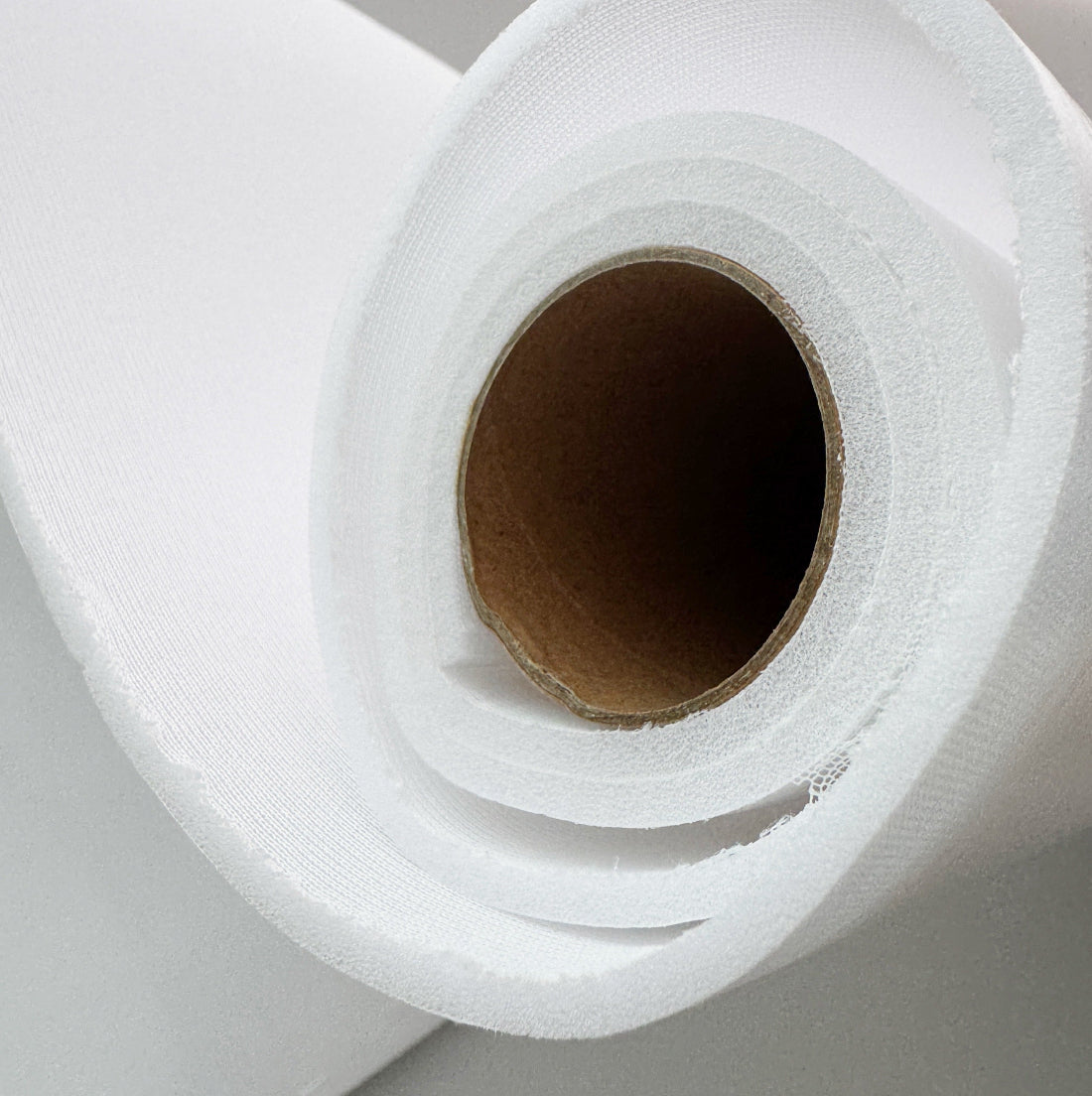 A close-up of a white roll of fabric or paper with a brown cardboard core, photographed against a light neutral background.
