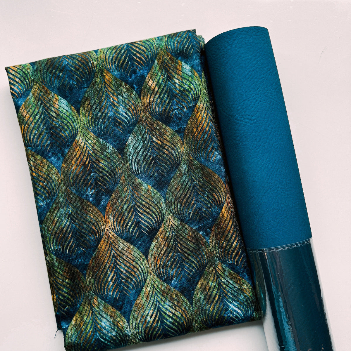 A folded piece of Tranquil Patina Horizon (Stock #48118 14) designed by Debbie Maddy for Moda Fabrics alongside a roll of solid teal faux leather.
