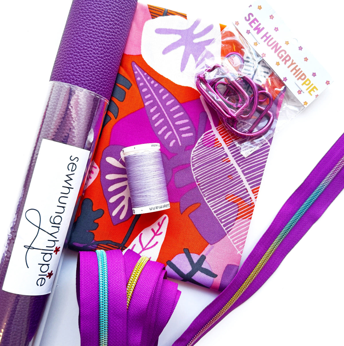 A flat lay of a Sew Hungry Hippie bag-making kit featuring a roll of purple faux leather vinyl, tropical leaf print fabric, matching purple hardware, dual-color zippers, and thread.
