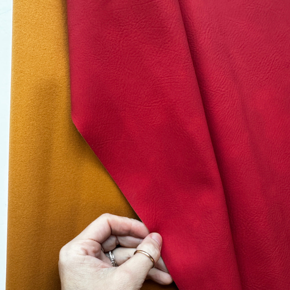 Close-up of red faux leather fabric with a subtle grain texture held by a hand over mustard-yellow textured fabric, showcasing contrasting materials and colors.