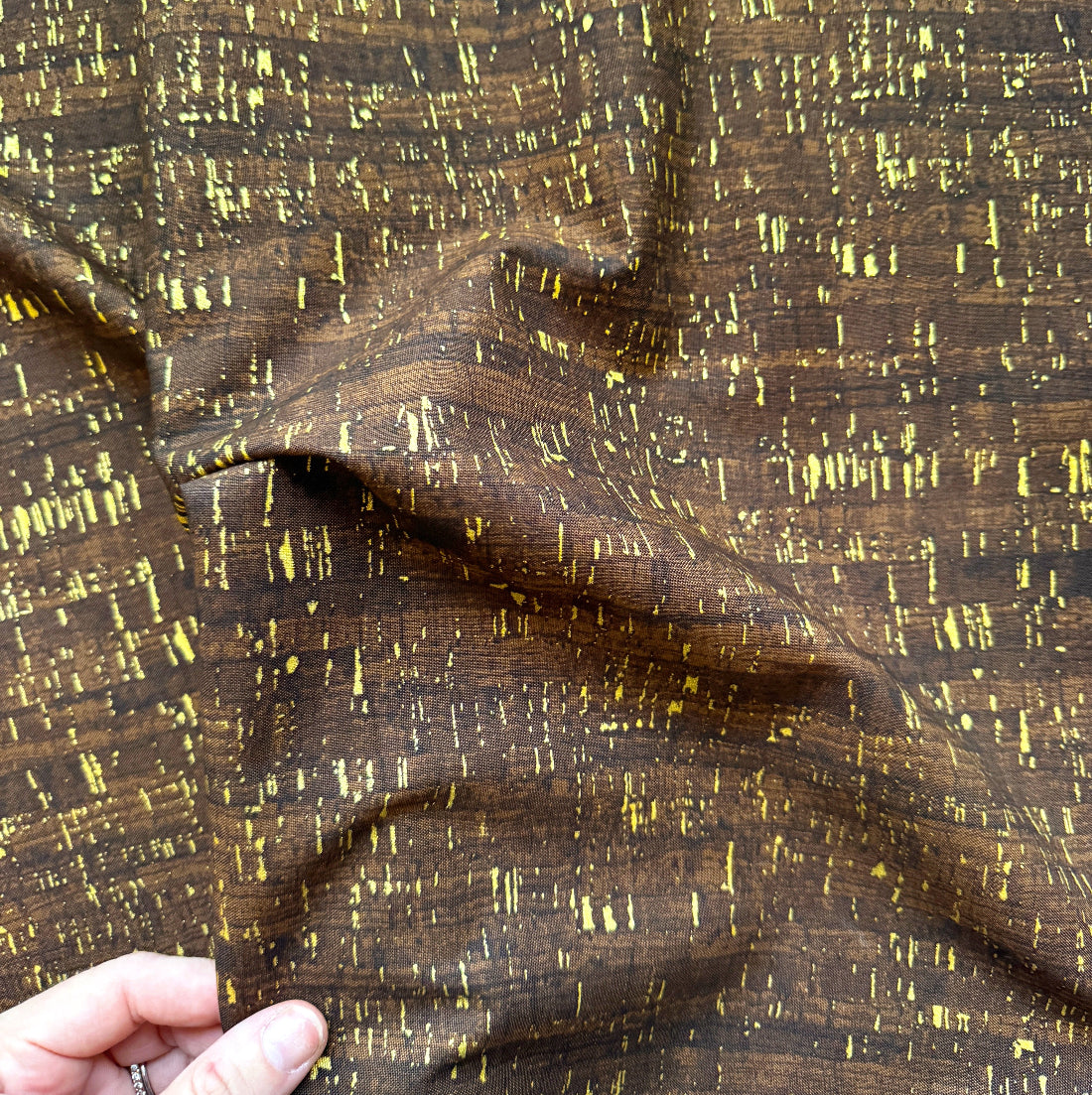 A close-up of dark brown fabric with a textured, distressed pattern of yellow and gold vertical streaks, held by a hand in the bottom-left corner.