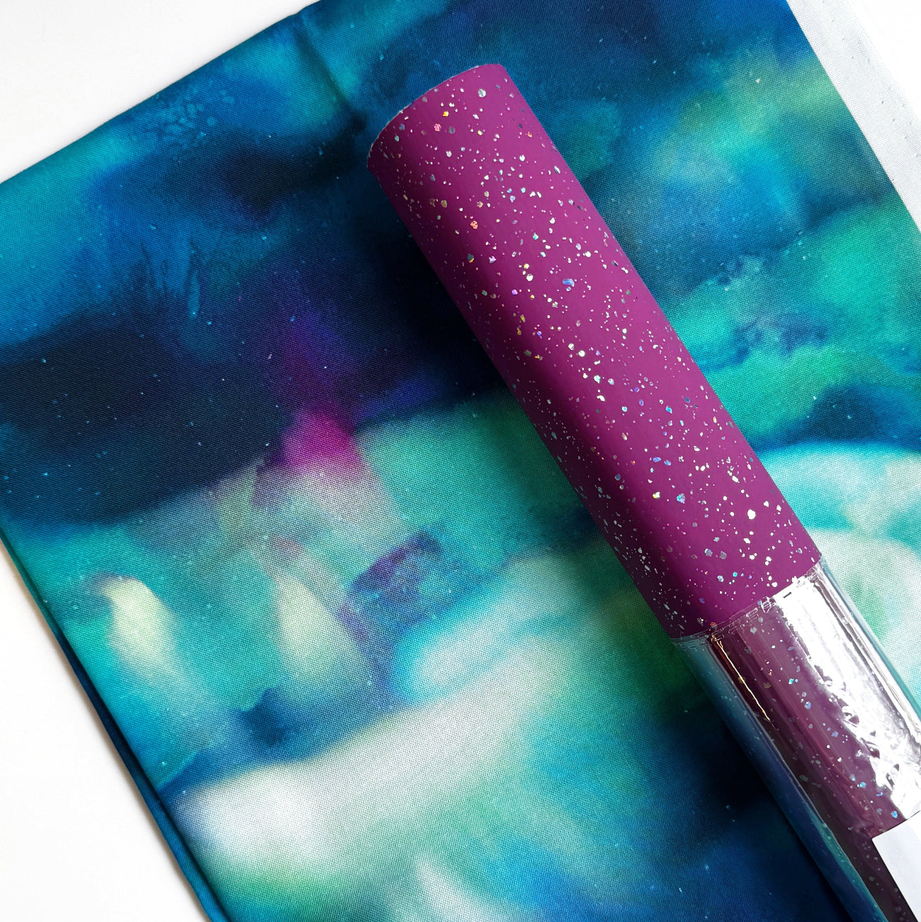 A roll of magenta purple glitter vinyl sheet placed over a piece of folded aurora-style watercolor fabric in shades of teal, blue, and green.