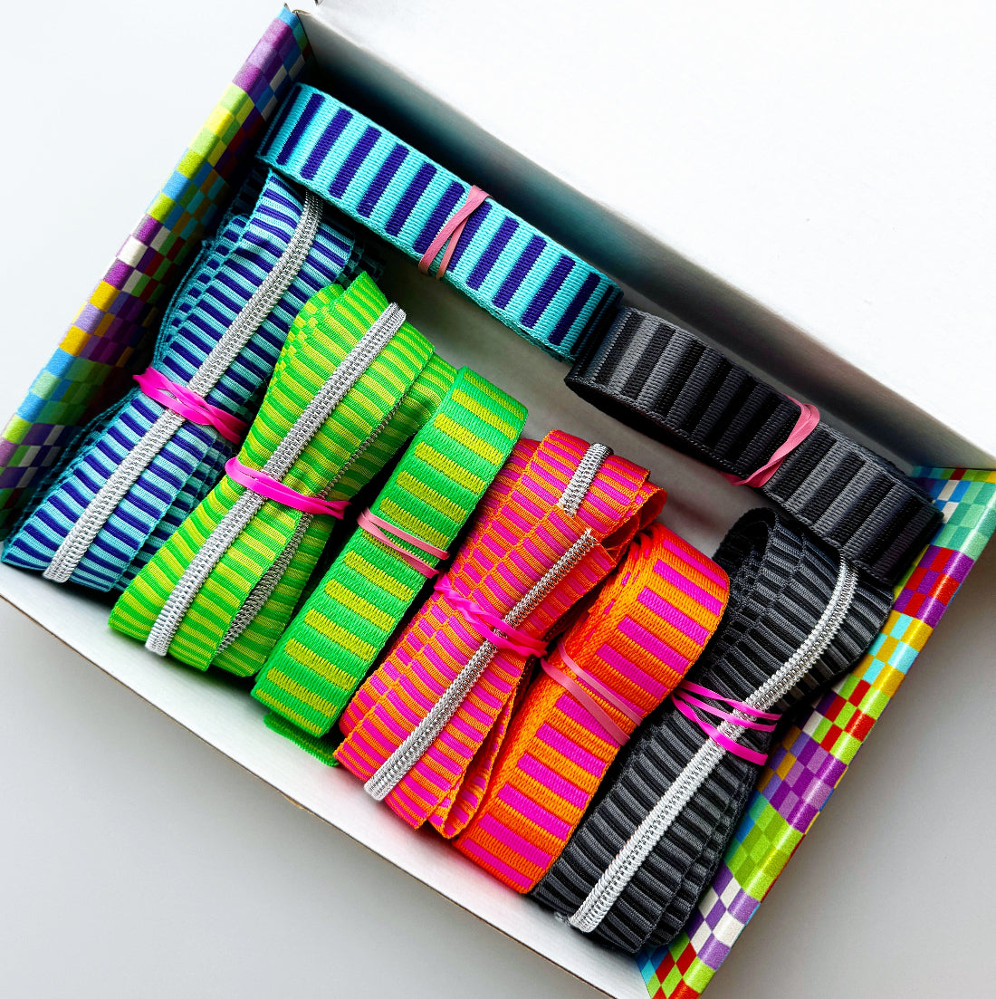 A box of colorful striped elastic bands or straps, arranged diagonally. The bands come in multiple vibrant colors including blue, green, pink, orange, and black, each tied with a small pink elastic band, set against a clean white background.
