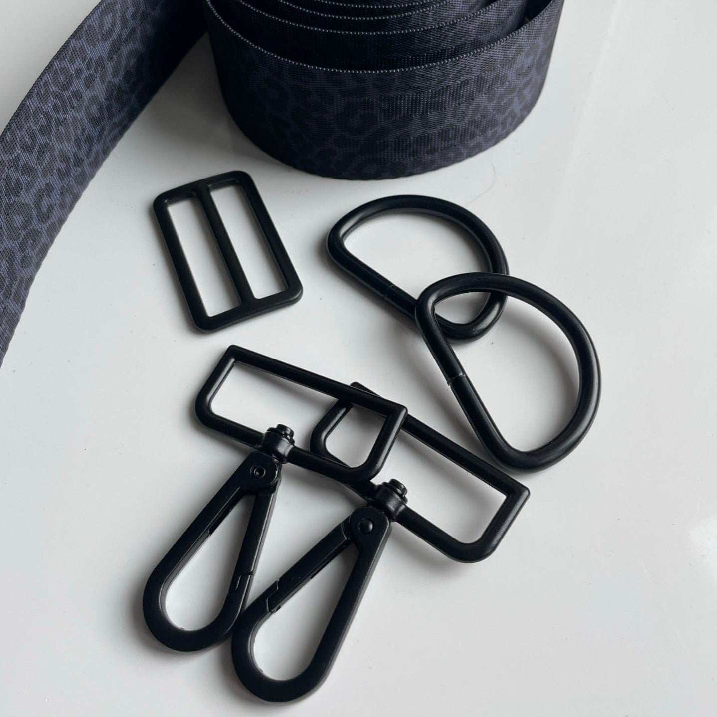 A black-and-grey leopard printed strap sits coiled next to two D-rings, two swivel hooks, and one adjustable buckle.