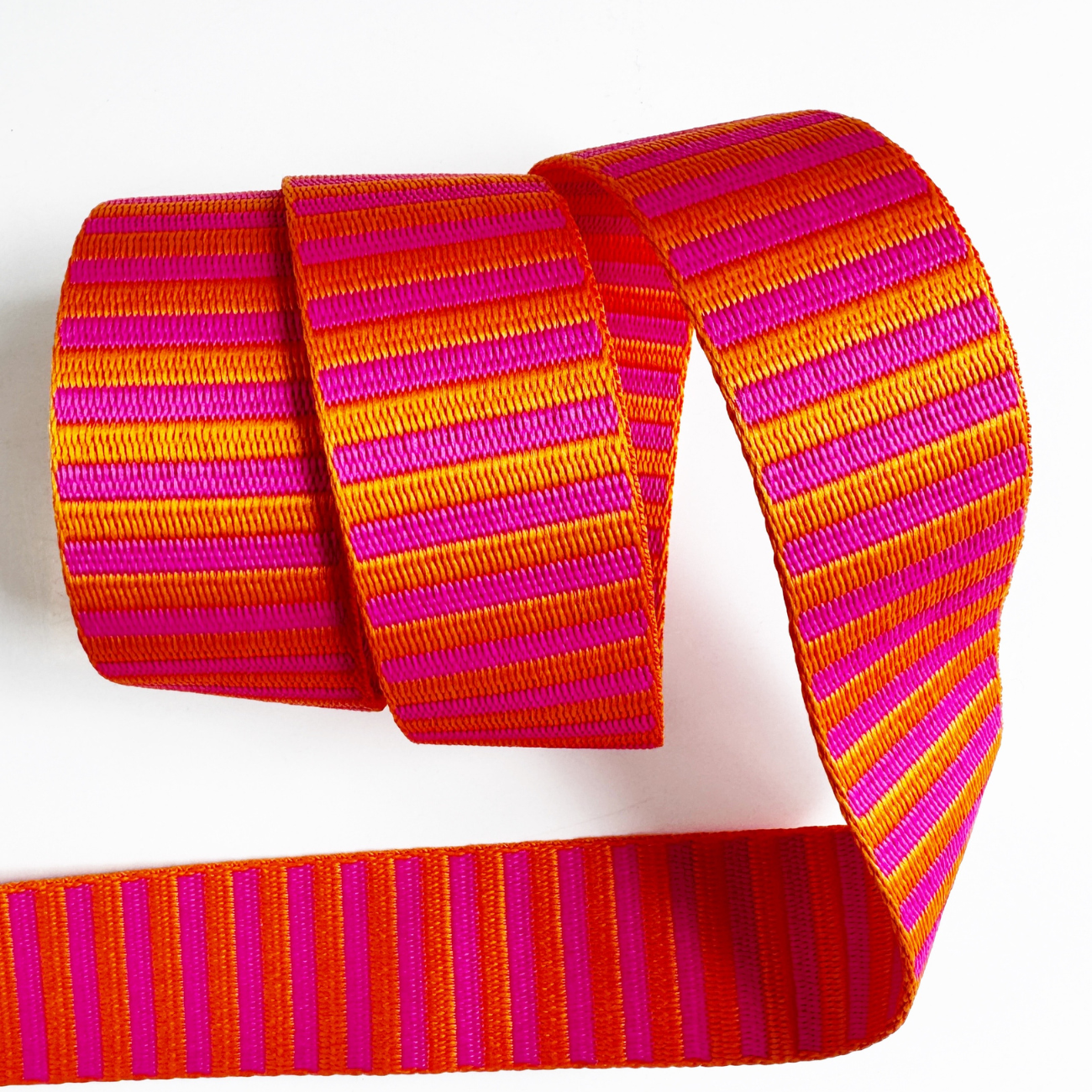 A 1.5-inch wide jacquard woven webbing strap showcasing a vibrant hot pink and orange horizontal stripe pattern, loosely coiled against a white background.