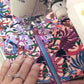 A close-up of a sewing machine stitching a colorful floral fabric panel with a purple zipper. A hand with manicured nails and rings guides the fabric.