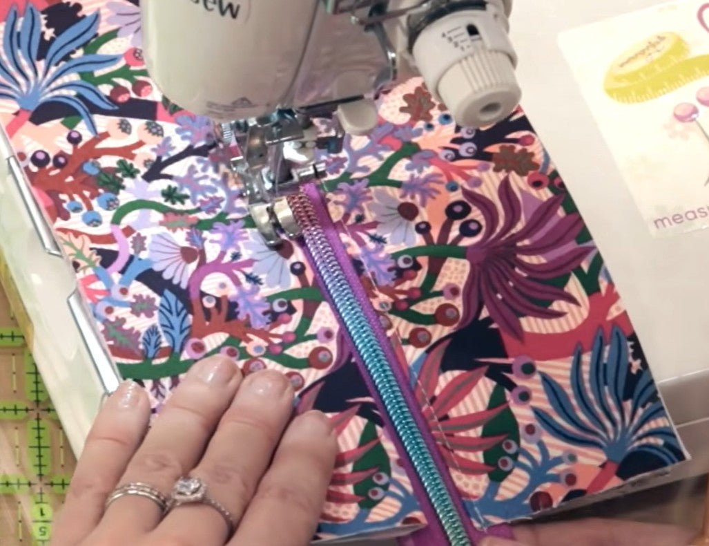 A close-up of a sewing machine stitching a colorful floral fabric panel with a purple zipper. A hand with manicured nails and rings guides the fabric.