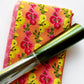 A piece of two yards folded orange fabric featuring a vibrant pattern of yellow, pink, and red flowers with green stems, resting next to a pink patterned fabric strip and a shiny roll of metallic green and silver foil.