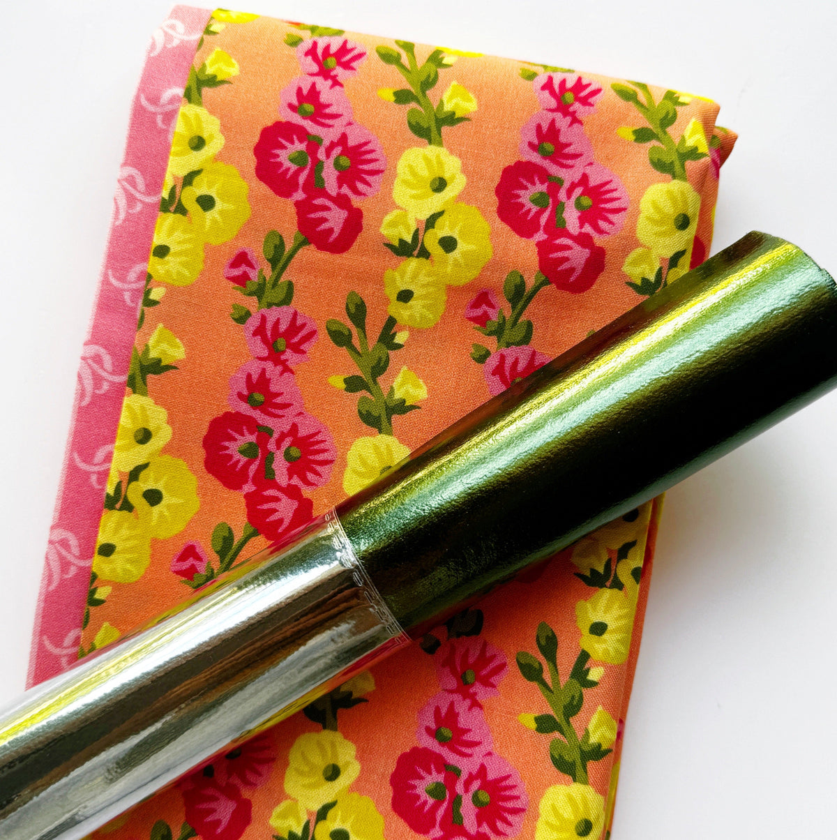 A piece of two yards folded orange fabric featuring a vibrant pattern of yellow, pink, and red flowers with green stems, resting next to a pink patterned fabric strip and a shiny roll of metallic green and silver foil.