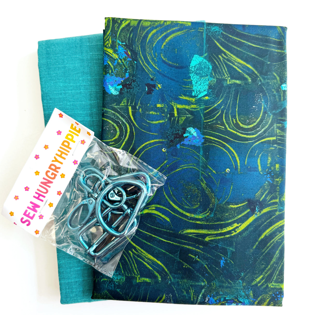 A craft bundle containing two yards of fabric, including a solid teal textile and a patterned blue and green abstract print, paired with a pack of matching metallic blue hardware clips from Sew Hungry Hippie.