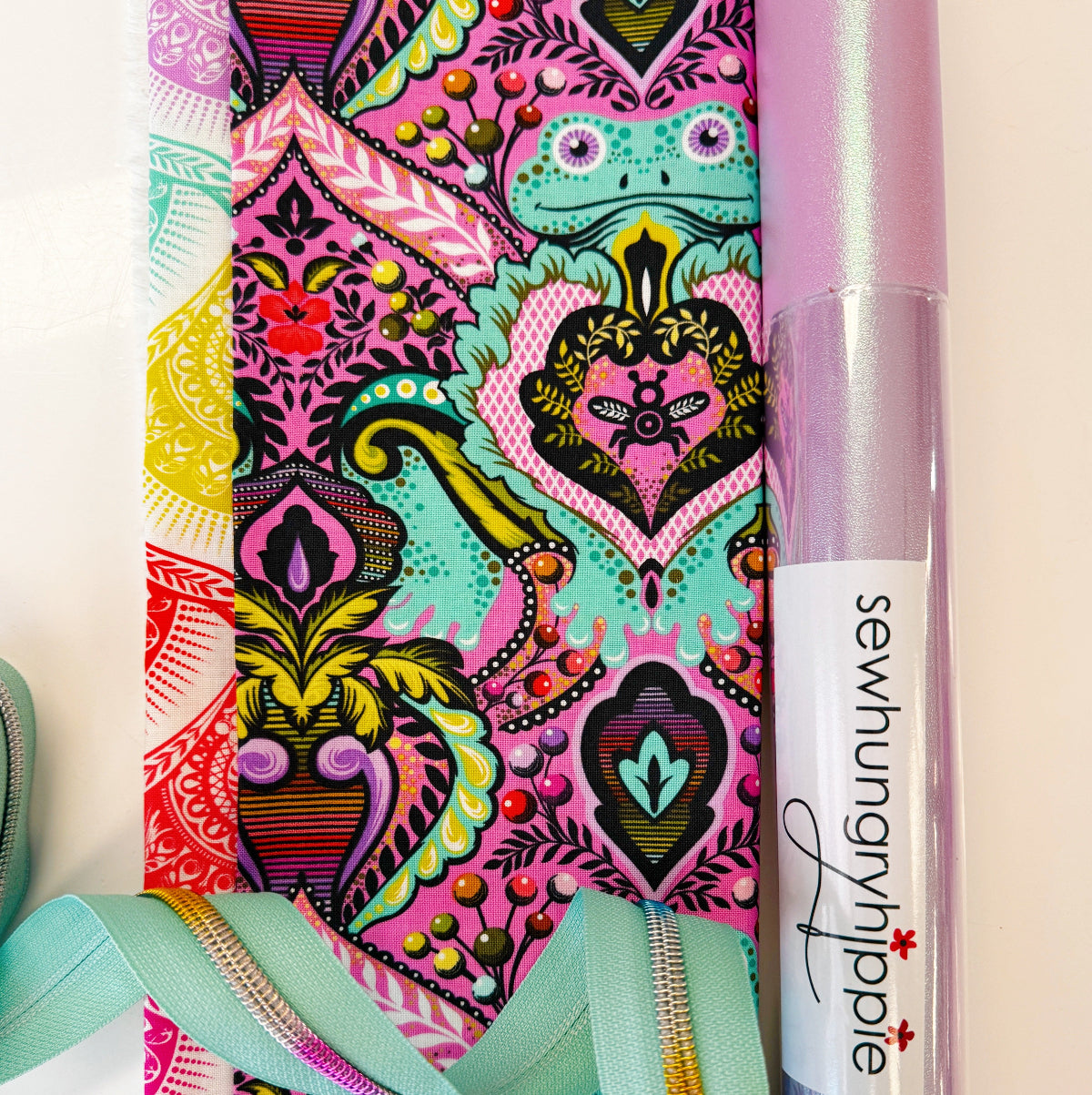 A close-up of a colorful patterned fabric or ribbon roll with bright pink, teal, yellow, and red ornamental motifs, alongside a tube of iridescent lavender sewing thread and a mint green ribbon on a light background.