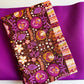 An overhead view of crafting fabrics featuring the "Khokhloma Soul" fabric from the Soul Fusion collection by Art Gallery Fabrics. The folded piece of fabric showcases vibrant purple and orange botanical prints on a dark brown textured background with a striped decorative border. It rests on top of a smooth sheet of solid, magenta-purple bonded nylon material.