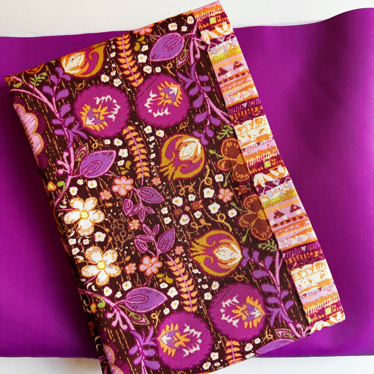 An overhead view of crafting fabrics featuring the "Khokhloma Soul" fabric from the Soul Fusion collection by Art Gallery Fabrics. The folded piece of fabric showcases vibrant purple and orange botanical prints on a dark brown textured background with a striped decorative border. It rests on top of a smooth sheet of solid, magenta-purple bonded nylon material.