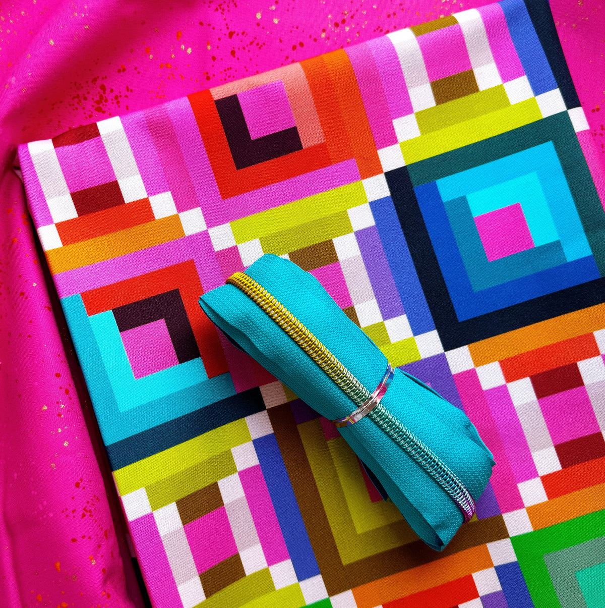 Fuschia pink speckled fabric in the background and a yard folded of geometric Happy Place collection by Annabel Wrigley for Windham Fabrics and coordinating turquoise zipper tape in rainbow teeth.