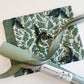 A bag making supplies kit featuring curated materials from Sew Hungry Hippie. The bundle includes a folded piece of Dragon Lair Sage fabric with  Ruby Star Society Fire Glow "Dragon Lair" (Watercress colorway) – 100% Premium Cotton Canvas teal and gold metallic dragon print, a roll of sage green faux leather vinyl fabric with a pebbled texture, and a length of matching sage green nylon coil zipper tape with metallic teeth drape across the top.