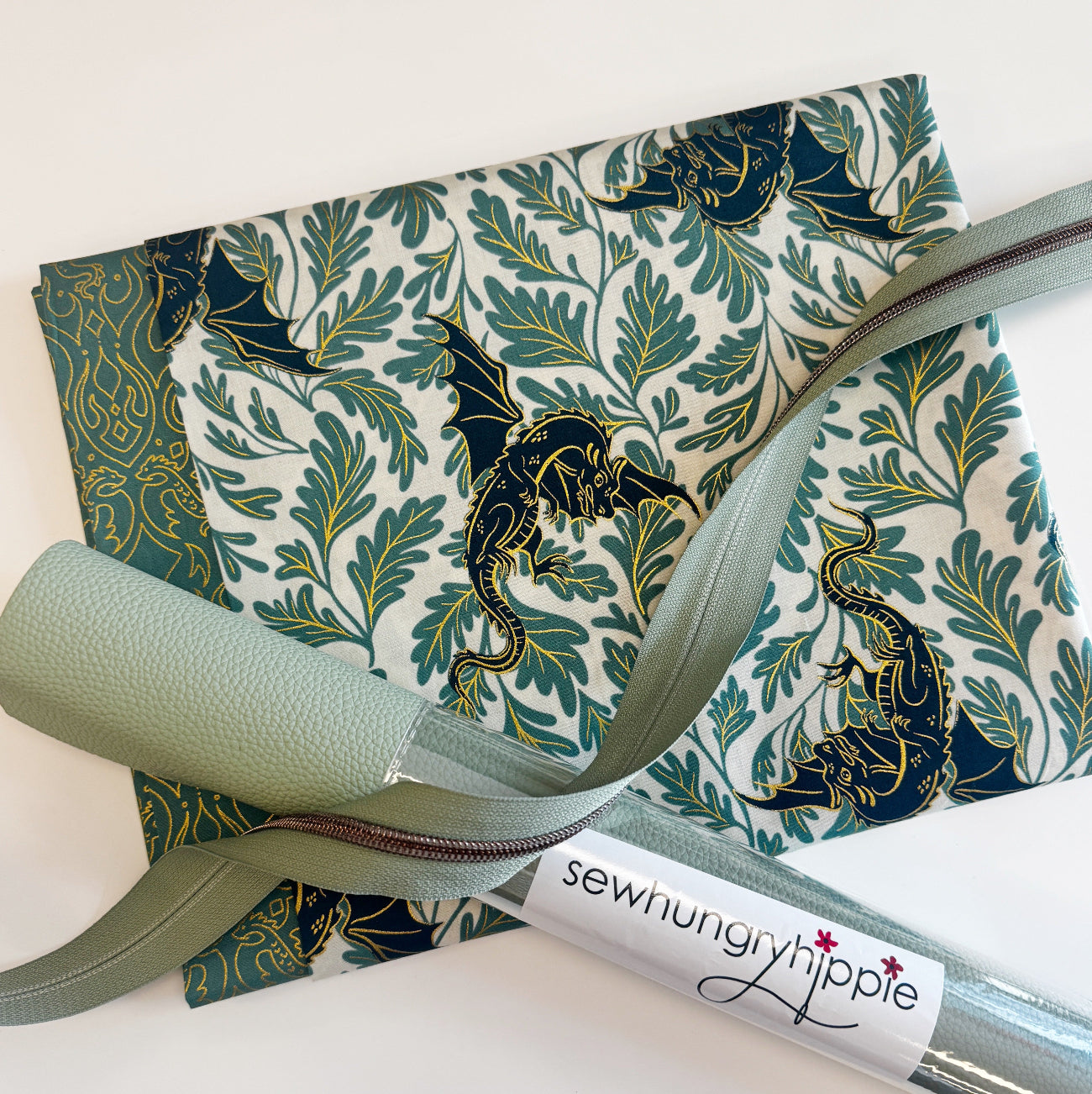 A bag making supplies kit featuring curated materials from Sew Hungry Hippie. The bundle includes a folded piece of Dragon Lair Sage fabric with  Ruby Star Society Fire Glow "Dragon Lair" (Watercress colorway) – 100% Premium Cotton Canvas teal and gold metallic dragon print, a roll of sage green faux leather vinyl fabric with a pebbled texture, and a length of matching sage green nylon coil zipper tape with metallic teeth drape across the top.
