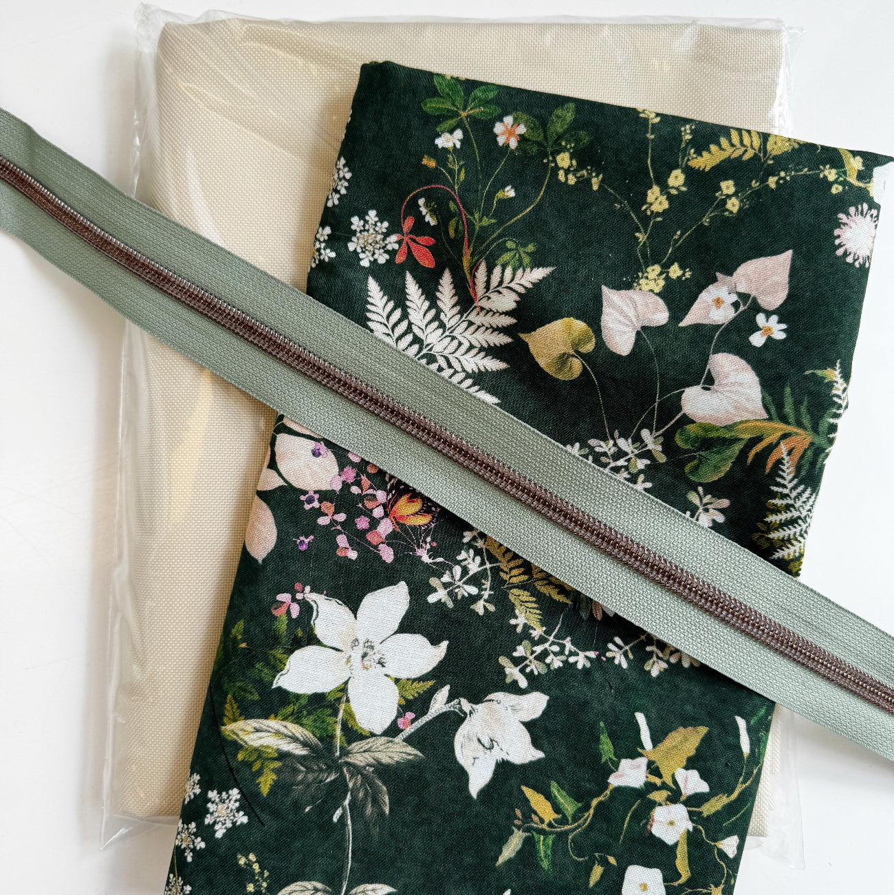 Dark forest green canvas fabric featuring a woodland wildflower botanical pattern, paired with a cream linen base fabric and a sage green nylon coil zipper tape for custom pouch and bag making.