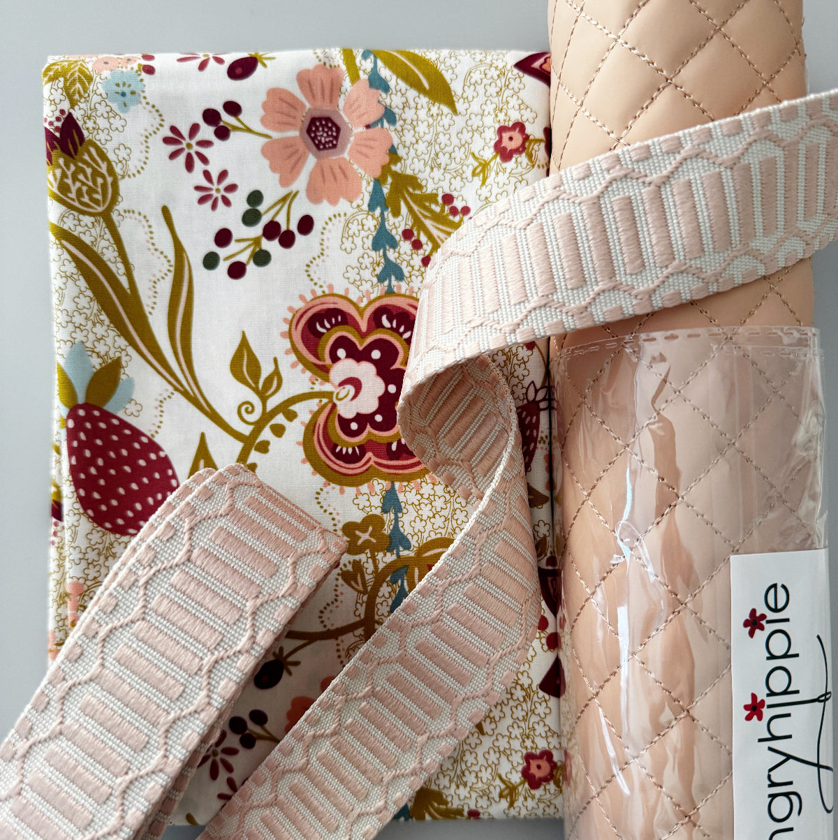 A close-up product shot showing a floral fabric item, likely a quilted blanket or throw, with a rolled section wrapped in a pale blush quilted fabric and a matching patterned woven ribbon. A visible label reads 'angryhippie'.