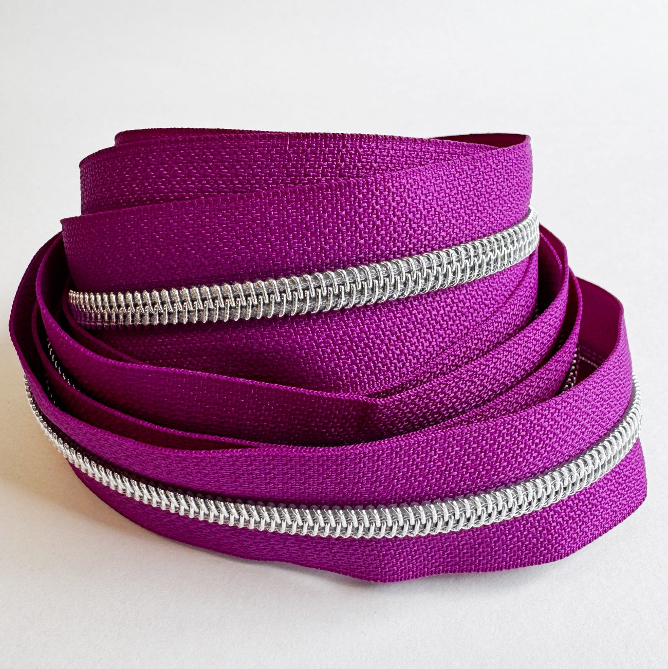 A length of teal nylon zipper tape loosely arranged into a circular, coiled nest shape on a clean white surface. The continuous rows of shiny silver zipper teeth track along the edges of the woven fabric, highlighting its flexibility.