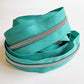 A length of teal nylon zipper tape loosely arranged into a circular, coiled nest shape on a clean white surface. The continuous rows of shiny silver zipper teeth track along the edges of the woven fabric, highlighting its flexibility.