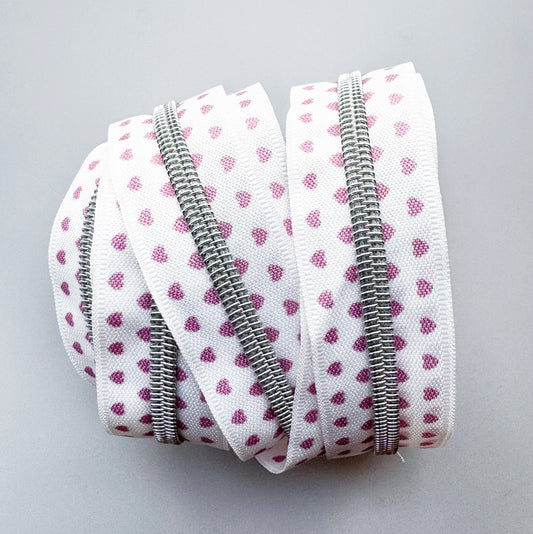 A coiled roll of white zipper tape trim featuring a pattern of small pink hearts, with a silver metal zipper running down the center, set against a plain light gray background.