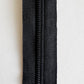 A close-up of a black metal zipper with a black fabric tape, shown vertically against a plain white background.