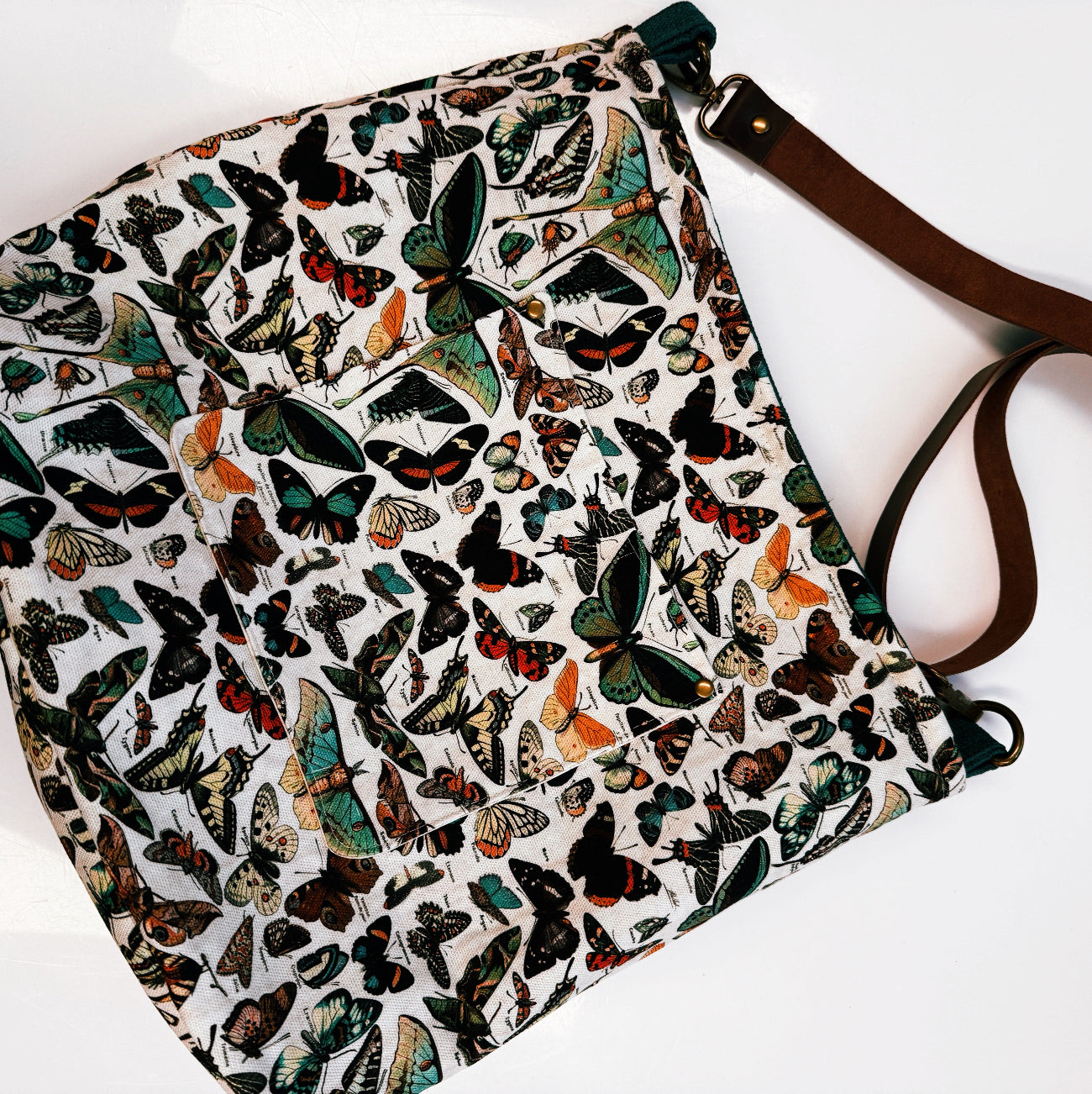 A fabric shoulder bag with an all-over vintage-style butterfly print on a white background, featuring a brown leather strap and a small front pocket.