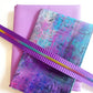 Grunge purple and teal abstract vintage script print fabric paired with a solid lavender canvas base and a purple striped rainbow teeth nylon coil zipper tape for custom pouch and bag making.