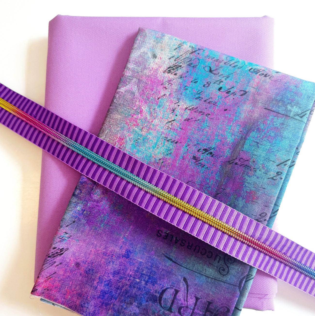 Grunge purple and teal abstract vintage script print fabric paired with a solid lavender canvas base and a purple striped rainbow teeth nylon coil zipper tape for custom pouch and bag making.