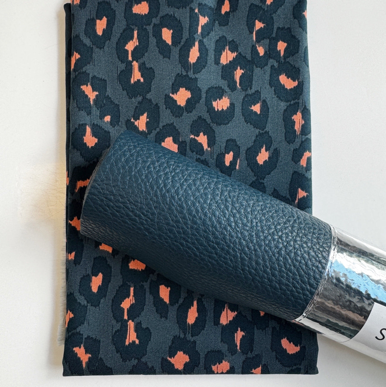 Charcoal gray and muted coral ikat leopard print cotton fabric paired with a coordinating dark teal pebbled faux leather roll for handmade bag making and sewing crafts.