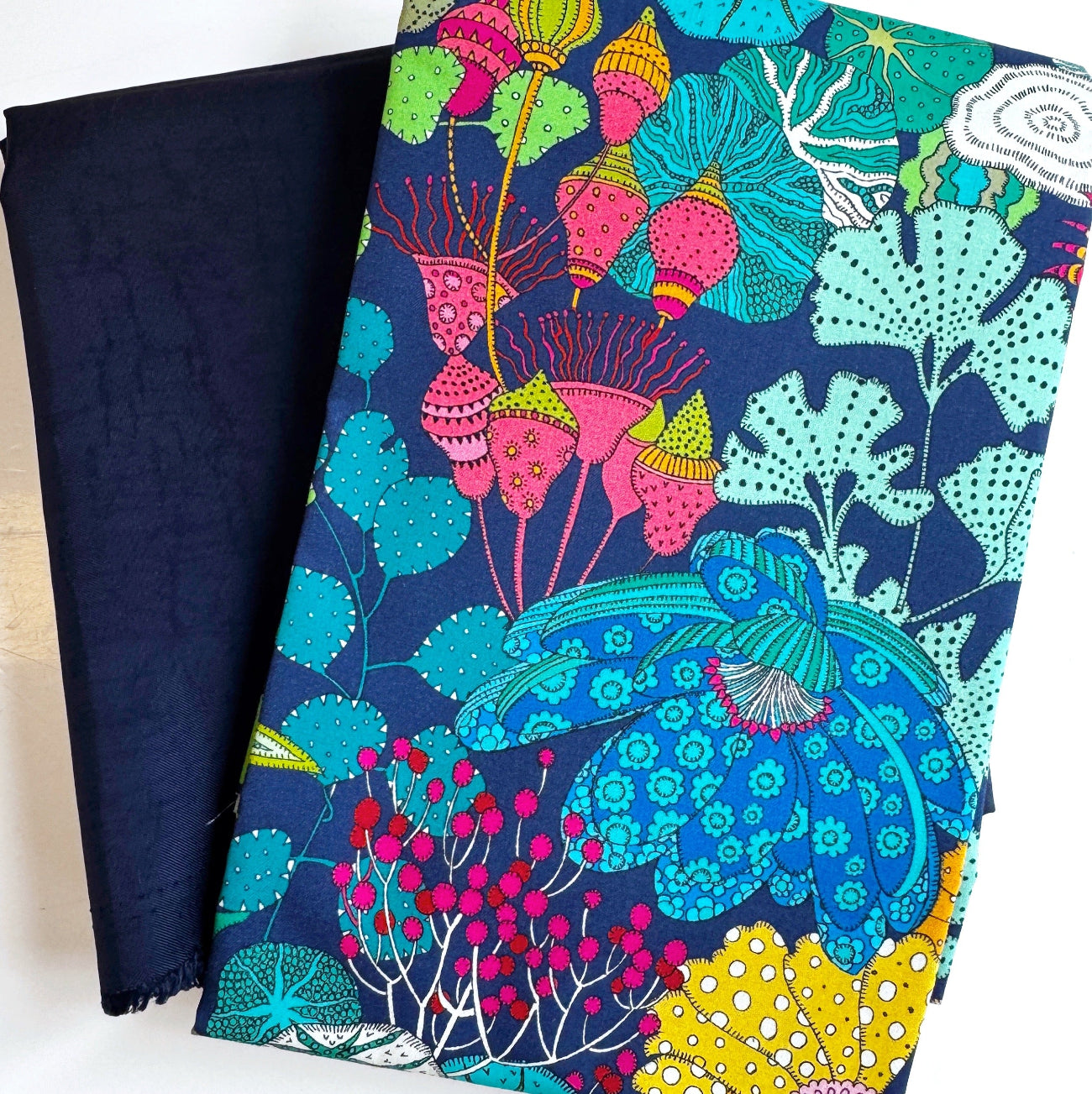 Vibrant multi-color whimsical botanical print fabric featuring bright blue flowers and lime green ginkgo leaves, paired with a solid dark navy blue canvas base fabric for sewing and bag making.