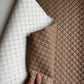A close-up of a quilted faux leather fabric shown in two colors: a warm taupe-brown and an off-white/cream. A hand is visible holding the corner of the cream piece to show its texture and thickness.
