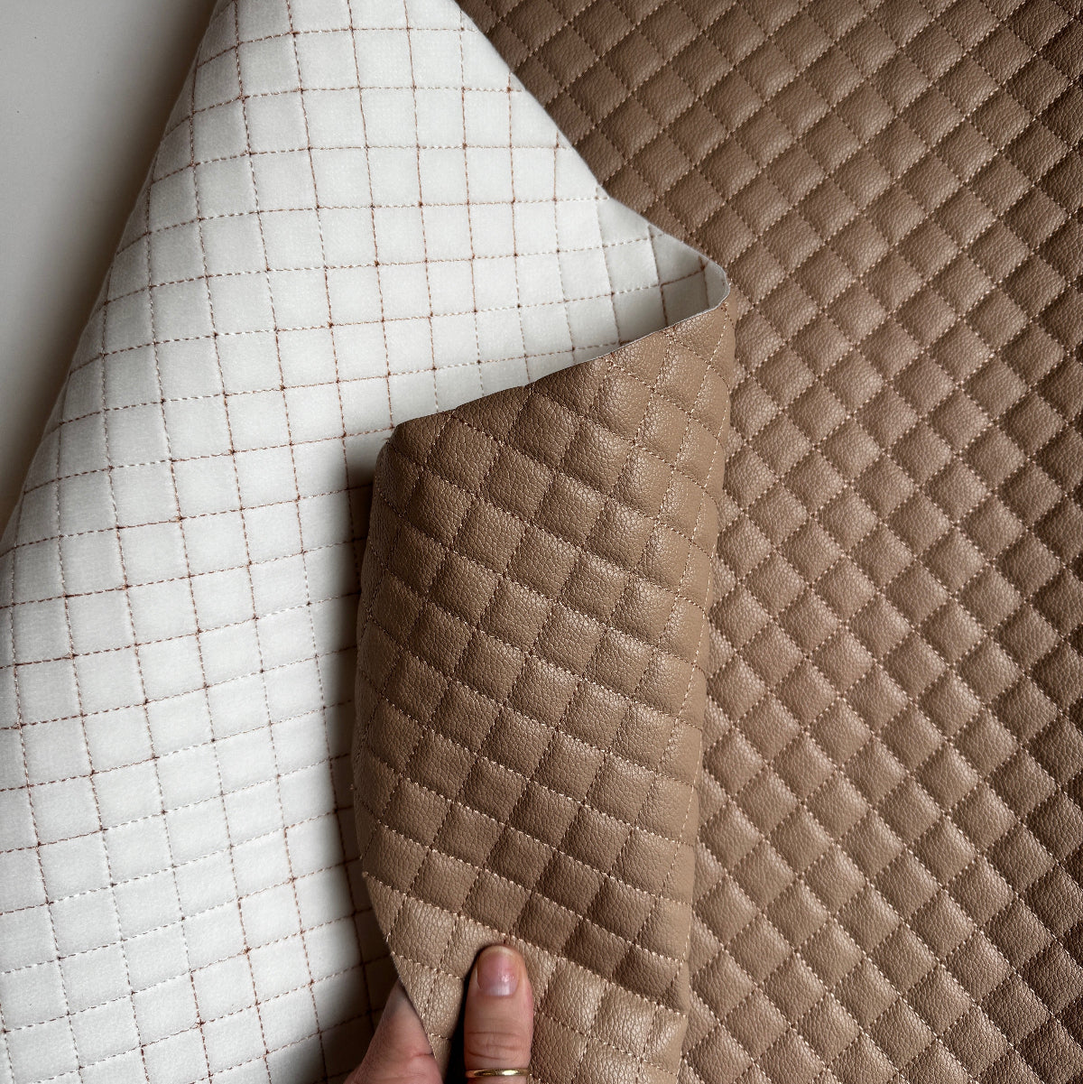 A close-up of a quilted faux leather fabric shown in two colors: a warm taupe-brown and an off-white/cream. A hand is visible holding the corner of the cream piece to show its texture and thickness.