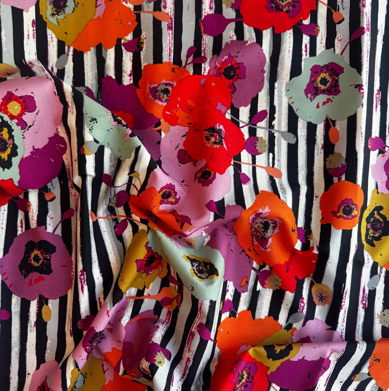 Art Gallery Fabrics Paparounes Spices softly draped. The design features a bold, artistic pattern of large watercolor-style poppy flowers in vibrant red, orange, plum purple, mustard yellow, and sage green scattered over a distressed black and white vertical striped background. 