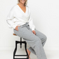 Woman sitting on a stool wearing a white robe and gray pants against a white background