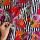 A close-up shot of a hand holding a piece of Art Gallery Fabrics Paparounes Spices premium floral stripe cotton fabric, showing the scale of the large poppy print and fabric drape. The design displays painterly flowers in rich spice tones of burnt orange, bright red, magenta, and olive yellow over a black and white vertical paint-stroke stripe pattern.