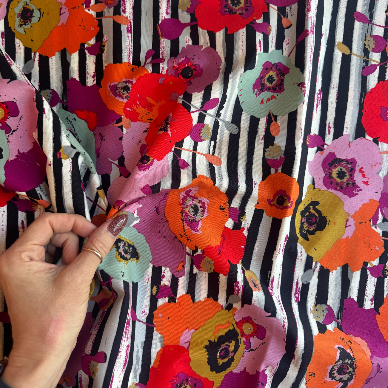 A close-up shot of a hand holding a piece of Art Gallery Fabrics Paparounes Spices premium floral stripe cotton fabric, showing the scale of the large poppy print and fabric drape. The design displays painterly flowers in rich spice tones of burnt orange, bright red, magenta, and olive yellow over a black and white vertical paint-stroke stripe pattern.