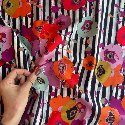 A close-up shot of a hand holding a piece of Art Gallery Fabrics Paparounes Spices premium floral stripe cotton fabric, showing the scale of the large poppy print and fabric drape. The design displays painterly flowers in rich spice tones of burnt orange, bright red, magenta, and olive yellow over a black and white vertical paint-stroke stripe pattern.