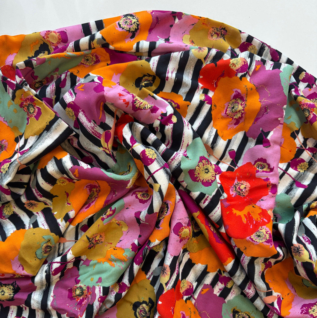 Spice Fusion Stripes rayon fabric by Art Gallery Fabrics featuring large orange, pink, and mint poppies layered over vertical black and white stripes. This soft, 100% rayon fabric has a fluid, silky drape ideal for sewing garments like dresses, tunics, and pants.